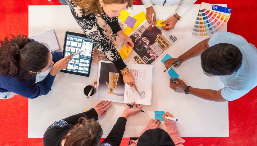 A group of students sit around a table, pointing at sketchbooks, colour swatches and digital design references