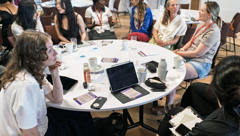 A group of diverse students seated around a circular table, engaged in conversation during a meeting or workshop, with laptops, tablets, and beverages on the table.